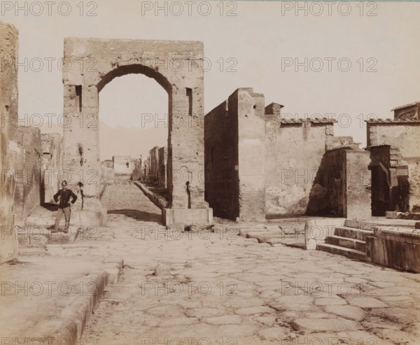 Strada Mercurio, View of the Strada Mercurio in Pompeii, Italy, Pompei, photograph, number: '1255.', collector's mark: 'VOSMAER', photographer: Giorgio Sommer, Pompeii, 1857 - 1875, Giorgio Sommer, photographer, pasteboard, paper, albumen print, height 315 mm x width 420 mm, street, ruin of a building, architecture, landscape with ruins, gate, entrance, A wide cobbled street stretches toward a large brick archway with rough, weathered surfaces and rectangular openings, flanked by high stone and plaster walls showing patches of wear and irregular edges; low stepped curbs and worn stone slabs line the foreground and a set of stone steps appears to the right, while on the left a uniformed man stands with hands on hips on a raised stone block; beyond the arch the street continues between rows of ruins and low buildings with tiled roof edges and uneven facades, and small stone markers sit near the base of the arch.