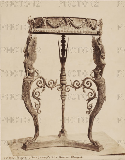 Tripod from the temple of Isis in Pompeii, Italy, Trepied (Oura) temple d'Isis: bronze: Pompei, photograph, number: '54', number: 'No. 5695', photographer: attributed to Giorgio Sommer, Pompeii, 1857 - 1914, Giorgio Sommer, photographer, Attributed to, pasteboard, paper, albumen print, height 444 mm x width 331 mm, furniture and household effects, temple, shrine, non-Christian religions, Tempio di Iside, An ornate metal stand with a circular top decorated with garland motifs rests on three elaborately carved supports; two side supports feature winged human-animal figures with detailed feathered wings and seated feline bodies atop scrollwork arms, while the central support is a vertical shaft with carved ornamentation and a stylized silhouette at its upper section; the lower ends of the supports terminate in curved legs with pawed feet standing on a rectangular base, and the piece is shown against a plain light background.