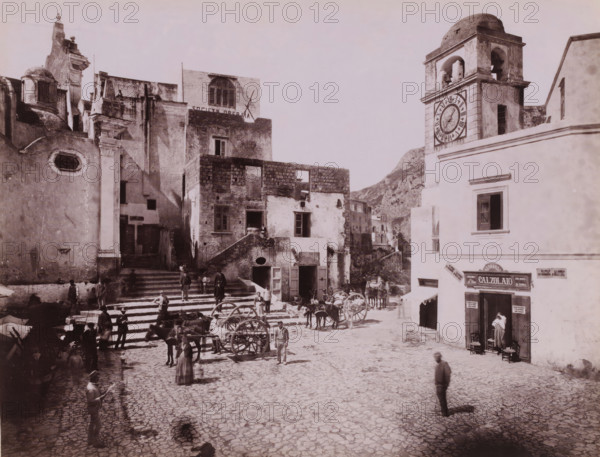 People on a square in Capri, Italy, Capri, photograph, inscription: 'Capri?', number: '6119.', photographer: attributed to Giorgio Sommer, 1857 - 1914, Attributed to, pasteboard, paper, albumen print, height 316 mm x width 445 mm, square, circus, Isola di, A sunlit town square with worn stone steps and a cobbled surface, flanked by multi-story buildings with weathered facades, arched windows, and crumbling plaster; a tall clock tower with visible bells rises on the right, and a row of shops occupies the ground floor beneath a prominent window; scattered carts with large wooden wheels and a few standing horses are positioned near the steps while people in period clothing stand, sit, and walk across the square, including a woman in a long dress near the foreground and several men clustered around the carts; narrow alleys and stairways lead up between the buildings, and a rocky hillside appears in the background.