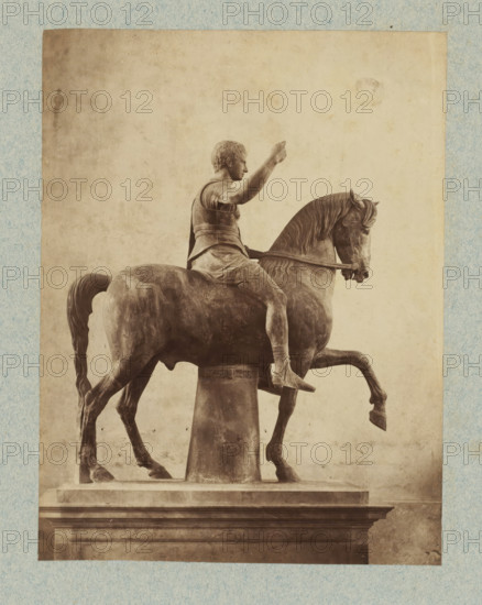 Equestrian statue, possibly of a Roman emperor, photograph, collector's mark: 'VOSMAER', Italy, after sculpture by anonymous, 1851 - 1890, photographer, after sculpture by, pasteboard, paper, albumen print, height 325 mm x width 249 mm, equestrian statue, A sepia-toned photograph of an equestrian statue showing a figure seated on a horse, the rider wearing a short tunic and cuirass with one arm raised and the other holding the reins, the horse depicted with a lifted foreleg and detailed mane and tail, the sculpture mounted on a cylindrical support above a rectangular pedestal, the image framed by a textured light-colored border.