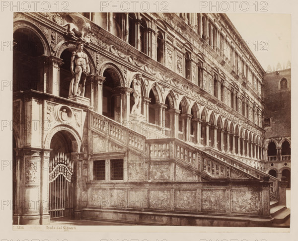 Scala dei Giganti at the Doge's Palace in Venice, Italy, Scala dei Giganti, Venezia, photograph, number: 'No. 3538.', inscription: 'Venetia Hof- Doge Paleis, Venetia', collector's mark: 'RIJKS SCHOOL FOR ARTS HANDBOOK, GROUP', number: '42', inscription: 'I', photographer: attributed to Giorgio Sommer, Venice, 1857 - 1914, Attributed to, pasteboard, paper, albumen print, height 320 mm x width 480 mm, palace, staircase, Doge's Palace, A sepia-toned photograph of an ornate stone staircase and arcade facade showing layered rows of arched openings and columns, decorative circular and rectangular relief panels, carved friezes and moldings, and balustraded stair flights. Two large nude male statues stand on pedestals at the top of the steps beneath archways, one holding an object at his side. A sculpted winged lion figure projects from an upper cornice. The staircase walls display panels of mottled stone and marble, and a decorative wrought gate is visible beneath an arch. The upper building ...