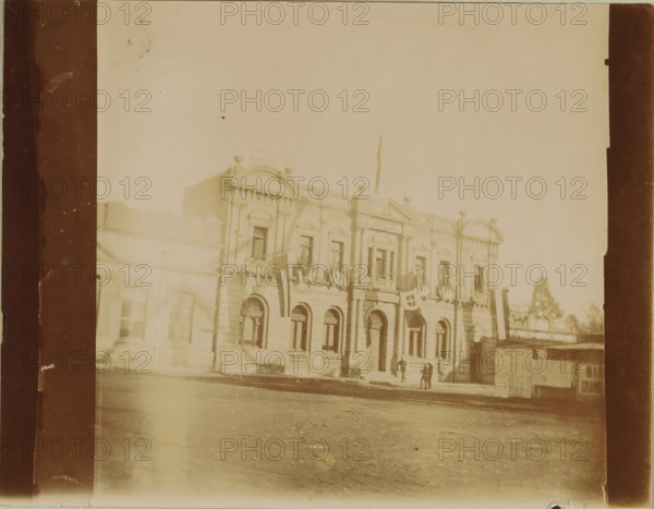 Exterior of a building, probably on Church Square in Pretoria, photograph, Pretoria, 1898, photographer, paper, baryta paper, daylight gelatin silver print, height 84 mm x width 108 mm, exterior, representation of a building, A sepia-toned photograph of a classical two-story stone building with arched windows on the lower level and rectangular windows above, featuring decorative pediments, columns, and balustrades along the roofline; flags and bunting hang from the facade and flagpoles rise above the roof; a few small groups of people stand near the main entrance steps; the foreground shows a paved or dirt street and the left and right edges of the image are framed by darker vertical borders.