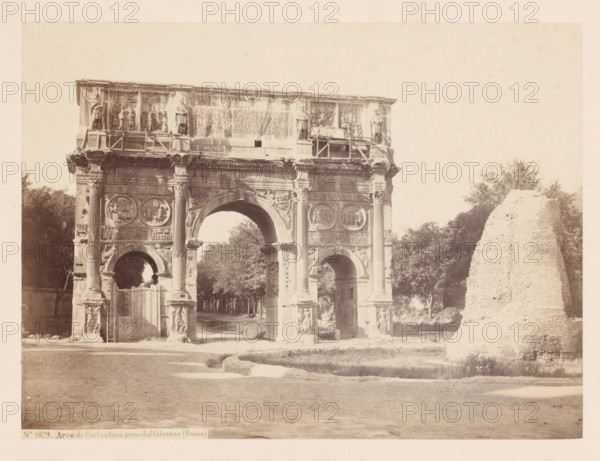 Arco di Costantino preso dal Colosseo (Roma), Triumphal Arch of Constantine in Rome, Italy, photograph, address: 'SOMMER & BEHLES, ROMA E NAPOLI', number: '8', number: 'No. 1029.', inscription: 'C. 85', inscription: 'Roma, A d Constantino', photographer: Sommer & Behles, Rome, 1867 - 1874, pasteboard, paper, albumen print, albumendruk, height 178 mm x width 240 mm, ruin of a building, architecture, triumphal arch, gate, entrance, Arch of Constantine, A stone triumphal arch with three arched passageways stands in the center, showing sculpted round reliefs, fluted columns, carved figures, and decorative friezes across its façade, with scaffolding and statues on the upper attic. Trees and a pathway are visible through the central arch and on either side of the structure. To the right of the arch there is a large, rough-textured ruined masonry mound. The foreground shows a paved road and a curved curb edge with a grassy strip beside it. The photograph has a pale, sepia tone and a wide bord...