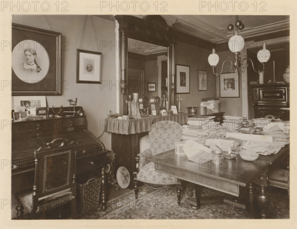 Study of Pieter Lodewijk Tak, photograph, photographer: Friedrich Carel Hisgen, Amsterdam, 1884 - 1907, paper, pasteboard, baryta paper, gelatin silver print, developed gelatin silver print, height 295 mm x width 361 mm, Person, Pieter Lodewijk Tak, interior of the house, A sepia-toned interior room filled with antique furniture and many stacks of papers on a large wooden table, showing ornate wooden chairs with patterned upholstery, a carved desk with a roll-top surface, and a wicker basket on the floor; framed portraits and pictures hang on the walls, including a large oval portrait of a woman, and smaller rectangular frames suspended by cords; a tall wooden mirror with decorative columns stands behind a side table covered with a draped cloth holding candlesticks, small framed photos, and decorative objects; a multi-arm ceiling lamp with frosted glass globes hangs above the table, and a patterned rug covers the floor beneath the furniture.