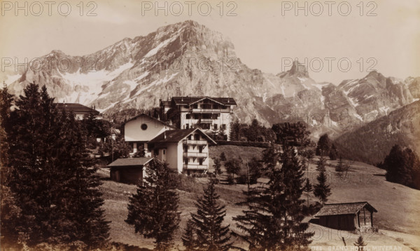 Grand Hotel Muveran, View of Grand Hotel Muveran at the foot of the Muveran in Switzerland, photograph, collector's mark: 'VOSMAER', number: '1786', Bernese Alps, 1900, photographer, Alps, paper, albumen print, height 133 mm x width 219 mm, hotel, hostelry, inn, mountains, Muveran, A mountainous landscape with steep rocky peaks and patches of snow rises in the background above a cluster of multi-story alpine buildings with pitched roofs and balconies; conifer trees and grassy slopes surround the buildings, with a small wooden barn visible on a hillside and scattered smaller trees and shrubs dotting the foreground.