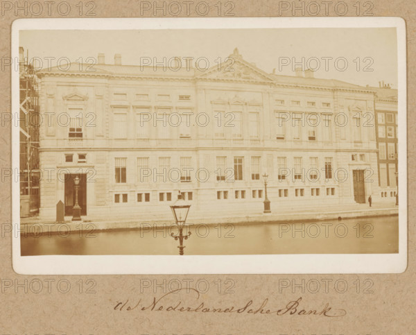 Façade of Oude Turfmarkt 127 in Amsterdam, former De Nederlandsche Bank, photograph, annotation: 'Nederlandsche Bank', Amsterdam, 1870 - 1890, photographer, paper, pasteboard, albumen print, height 94 mm x width 157 mm, height 162 mm x width 251 mm, bank, civic architecture, edifices, dwellings, Rokin, Oude Turfmarkt, A sepia-toned photograph of a large classical stone building with tall windows and decorative pediments along its facade, a triangular sculpted pediment above the central section, and chimneys visible on the roof; a canal runs in front of the building with a decorative street lamp reflected in the water, a second lamp visible on the sidewalk beside the building, scaffolding is present at the far left edge, and a solitary figure stands near a large doorway on the right side.