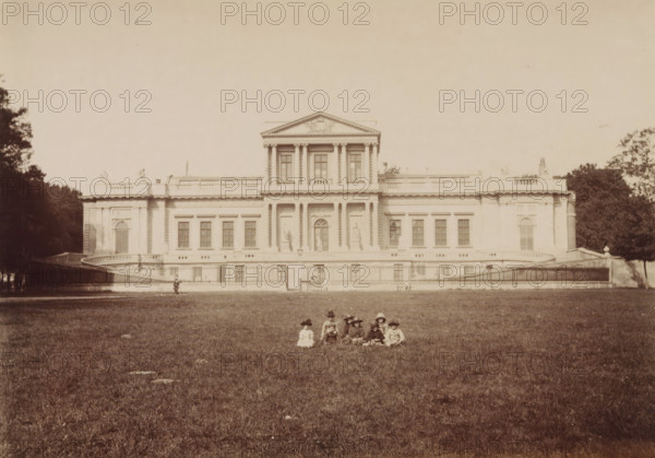 Paviljoen Welgelegen in Haarlem, with children sitting on the grass, photograph, annotation: '(top. K3: Haarlem I), A. Jager - also . Felix van West same persons, leporello H)', photographer: Andries Jager (possibly), Haarlem, 1870 - 1900, paper, pasteboard, albumen print, albumendruk, height 118 mm x width 167 mm, height 426 mm x width 301 mm, anonymous historical persons depicted in a group, in a group-portrait, civic architecture, edifices, dwellings, Paviljoen Welgelegen, A wide neoclassical building with a central pediment and columns stretches across the middle of the image, flanked by trees at the edges; a low wrought iron fence runs in front of the building above a grassy slope, and a group of children wearing period clothing and hats sit on the lawn in the foreground.