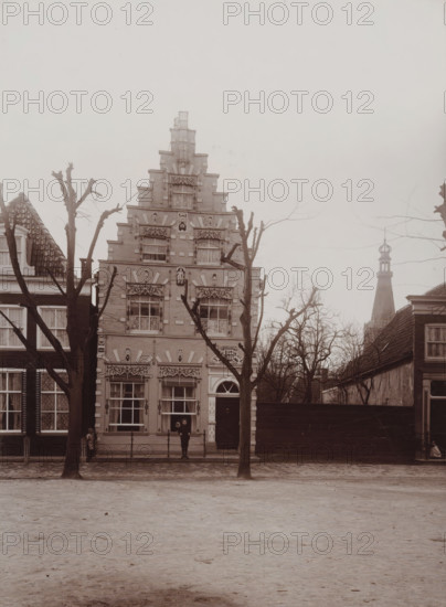 View of Radboud Castle in Medemblik, photograph, annotation: 'Medemblik, c.1900, 13 1, 2, Radboud Castle in Medemblik', inscription: 'B. Reekers, Photographer', photographer: Barend Reekers, Medemblik, 1890 - 1910, paper, pasteboard, daylight collodion silver print, height 160 mm x width 119 mm, height 132 mm x width 175 mm, landscape with tower or castle, Radboud Castle, A street scene showing a tall ornate brick building with a stepped gable and decorative window surrounds, flanked by adjacent buildings with tiled roofs, leafless pruned trees standing in front, a wooden gate or fence to the right, a distant tower with a pointed spire visible behind the trees, a cobbled or paved area in the foreground, and two small figures standing near the building entrance while another small figure is seated near the left side of the frame.