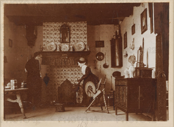 Interior of a living room, furnished in the old style of Texel with dolls, photograph, annotation: 'Old Texel room, Texel', annotation: '16 1, 2', Texel, 1890 - 1910, photographer, paper, pasteboard, daylight collodion silver print, height 117 mm x width 164 mm, height 220 mm x width 270 mm, interior of the house, A sepia-toned interior scene shows two people near a large fireplace with patterned tiles and decorative plates on the mantel; one person stands to the left wearing dark clothing and holding a long thin object, while the other, dressed in lighter clothing with a bonnet, leans over a spinning wheel in the center foreground; the room contains wooden furniture including a carved cabinet on the right, a small table on the left with stacked containers, wall-hung objects and framed pictures, various hanging metal utensils and a tall cylindrical stove or pipe, and the floor is wooden planks.
