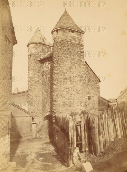 View of the Helpoort in Maastricht, photograph, inscription: 'Helpoort Maastricht', name: 'S. FRANCO, bi The Hague.', photographer: Héliodore Dandoy, Maastricht, c. 1875 - in or before 1884, paper, pasteboard, albumen print, height 222 mm x width 165 mm, city-gate, façade (of house or building), gate, entrance, Helpoort, A sepia-toned photograph of a stone building with two conical-roofed cylindrical towers, rough stone walls, and small window openings, flanked by brick structures on the left and a wooden palisade fence on the right, with a narrow dirt pathway leading toward an arched entrance at the base of the central tower and shadowed areas along the ground.
