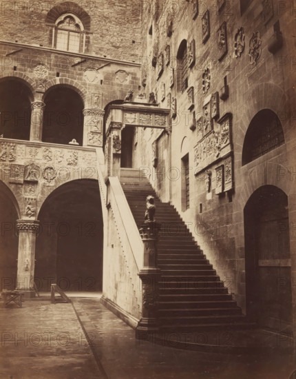 Stairs and courtyard of the Palazzo del Bargello in Florence, photograph, Florence, c. 1875 - c. 1900, photographer, Tuscany, paper, pasteboard, albumen print, albumendruk, height 250 mm x width 195 mm, courtyard, palace, staircase, A sepia photograph of a stone courtyard featuring a wide staircase with a solid balustrade leading upward to an ornate covered walkway, a sculpted lion figure atop a newel at the base of the stairs, and a series of arched openings and columns with carved capitals along the left side; the surrounding walls display numerous decorative relief panels and sculpted medallions, with a large arched doorway visible on the right and a tall window with a rounded top set high on the wall above the arches.