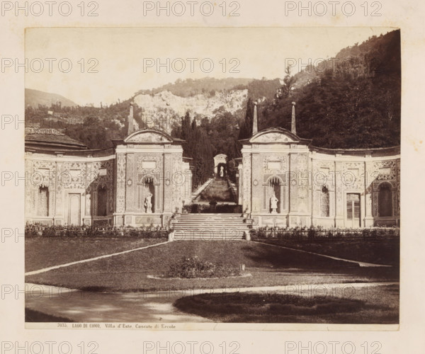 LAGO DI COMO. Villa d'Este, Cascate di ercole, Waterfalls in the garden of Villa d'Este on Lake Como, photograph, number: '7035.', Como, c. 1875 - c. 1900, photographer, Lombardy, paper, pasteboard, albumen print, height 202 mm x width 249 mm, water-works in garden, Cernobbio, Villa d'Este (Cernobbio), A wide sepia-toned photograph shows a formal building facade with ornate carved panels and archways flanking a central stairway that leads up to a terrace and a long narrow reflecting pool or canal framed by hedges and statues; tall slender obelisks topped with spherical finials rise above the roofline at intervals, and sculpted figures are placed in recessed niches on the facade and along the central axis; beyond the terrace a colonnaded pavilion or arch frames the distant end of the water feature, and dense tree-covered hills and a rocky cliff rise in the background, while a trimmed lawn and a curved path occupy the foreground.