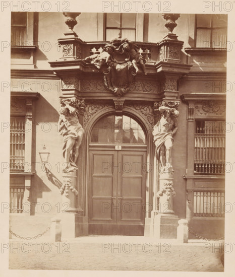 Fürstllich Liechtenstein'sches Palais, SEITEN-PORTAL', Side entrance of Stadtpalais Liechtenstein in Vienna, photograph, number: 'Nr. 48.', Vienna, publisher: Ludwig Niernberger, c. 1875 - c. 1900, photographer, Lower Austria, paper, pasteboard, albumen print, height 235 mm x width 197 mm, piece of sculpture, reproduction of a piece of sculpture, coat of arms (as symbol of the state, etc.), gate, entrance, A sepia-toned photograph of a building entrance with a large double wooden door set beneath an arched transom window, flanked by two tall sculpted male figures carved as supporting columns, ornate capitals, and decorative floral garlands on the column bases; above the arch is an elaborate carved entablature with scrollwork and a central cartouche topped by reclining cherub-like figures and decorative urns on pedestals; to either side of the entrance are windows with vertical bars and decorative moldings, a wall-mounted lantern with a bracket casting a shadow, short stone posts connec...