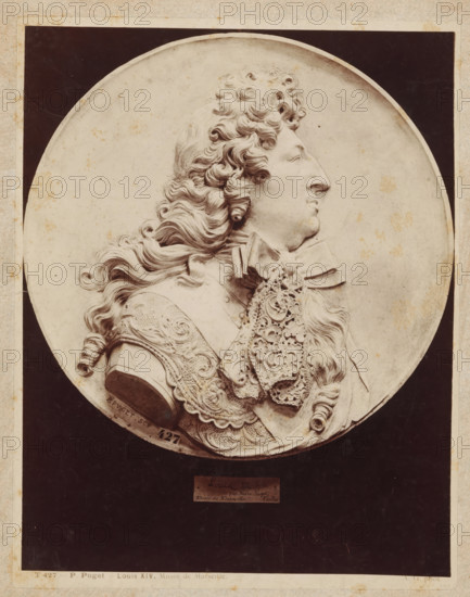 P. Puget Louis XIV. Musée de Marseille., Relief with profile portrait of King Louis XIV, by P. Puget, photograph, number: 'T 427', collector's mark: 'RIJKS-SCHOOL VOOR TEEKENONDERWIJZERS AMSTERDAM', photographer: Adolphe Giraudon, c. 1875 - c. 1900, Adolphe Giraudon, photographer, paper, pasteboard, albumen print, height 260 mm x width 204 mm, sculpture, historical persons ((full) bust portrait), sculpture (relief, sculpture), A circular relief portrait of a man in profile facing right set against a dark rectangular background, showing detailed curly hair cascading past his shoulder, a prominent nose and chin, and an ornate lace cravat and decorative armor with carved patterns and flourishes, all rendered in a monochrome stone or plaster tone with small specks and slight discolorations on the surface.