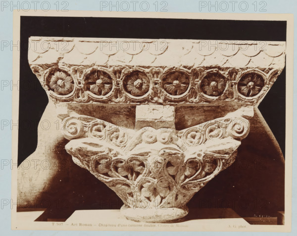 Capital of a double column with plant motifs, Art Roman - Chapiteau d'une colonne double. Cloître de Moissac., photograph, number: 'T 947', collector's mark: 'INSTITUUT VOOR KUNST-NIJVERHEIDSONDERWIJS GABRIËL METSUSTRAAT No. 16, AMSTERDAM', collector's mark: 'Dagteeken- en Kunstambacht- school voor Meisjes Amsterdam', name: 'A. GIRAUDON ÉDITEUR 15. R. BONAPARTE. PARIS', photographer: Adolphe Giraudon, publisher: Adolphe Giraudon, Paris, c. 1875 - c. 1900, Adolphe Giraudon, photographer, publisher, paper, pasteboard, albumen print, height 205 mm x width 260 mm, flowers, ornament, foliage, tendrils, branches, capital (, column, pillar), sculpture (relief, sculpture), A close-up photograph of an ornate carved stone capital showing a row of circular floral motifs along the top band, each set within a carved roundel, beneath a plain molded abacus with scalloped detailing; below the band a central rectangular block sits above a triangular arrangement of carved foliage and spiral volutes with...