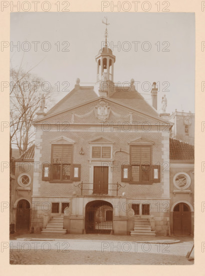 Middelharnis Town Hall, photograph, annotation: 'Town Hall, after the restoration 1909, Middelharnis', annotation: 'C. van Rhijn, 1683-1745', Middelharnis, 1909, photographer, Attributed to, paper, pasteboard, daylight collodion silver print, height 225 mm x width 163 mm, town hall, façade (of house or building), A symmetrical brick building facade with a triangular pediment topped by a small cupola and a decorative weather vane, flanked by statues on the roof corners and a tree visible to one side; shuttered windows with patterned panels and circular windows set into stone sections at either side; a central doorway set above a short staircase and balcony with wrought iron railing, an arched gated passageway beneath, and paired stone staircases leading to raised entrances; cobblestone street in front and a tall chimney and clock tower partially visible beyond one roofline.