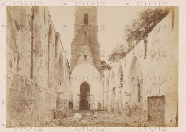 Interior of the Reformed Church in Middelharnis after the fire, photograph, annotation: 'Church of the Dutch Reformed Congregation on July 5, 1904, viewed towards the West, Middelharnis', annotation: 'Middelharnis (ZH), Church of the Dutch Reformed Congregation on July 5, viewed towards the west', Middelharnis, photographer, Attributed to, paper, pasteboard, albumen print, height 164 mm x width 231 mm, interior of church, A monochrome view of a ruined vaulted interior with high flanking walls and a central tower at the far end, showing weathered plaster and exposed brick surfaces, arched openings along both sides with some remaining columns and capitals, a tall pointed arch at the center beneath the tower with a recessed doorway and a barred window above, rubble and debris scattered across the floor, rough textured wall surfaces with patches of peeling material, and trees visible above the right wall outside the structure.