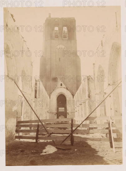 Interior of the Reformed Church in Monster after the fire, photograph, annotation: 'Monster NH Church (ZH), view to the west after the fire July 1901', annotation: 'NH Church, viewed to the West, after the fire, July 1901, Monster', Monster (South Holland), 1901, photographer, Attributed to, paper, pasteboard, albumen print, height 230 mm x width 170 mm, interior of church, A tall brick tower with large arched openings stands at the center of the image, flanked by the ruined walls of a long building with rows of pointed arch openings on both sides; the interior foreground shows wooden beams and planks laid across the space with angled support poles crossing toward the viewer, rubble and uneven ground visible beneath, and light filtering through the empty archways toward the distant tower.