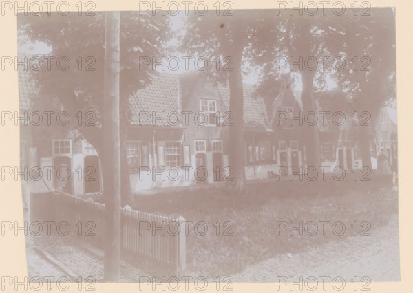 Courtyard of the Old Men's House in Naaldwijk, photograph, annotation: 'Facades of the Old Men's House to the west of the courtyard, May 1910, Naaldwijk', Naaldwijk, 1910, photographer, Attributed to, paper, pasteboard, daylight collodion silver print, height 157 mm x width 219 mm, courtyard, façade (of house or building), A row of brick houses with tiled gabled roofs is partially obscured by tall trees in front; each house has shuttered windows and small doorways, and some windows have visible windowboxes. A wooden picket fence runs along a grassy area in front of the houses, and a cobblestone path or street is visible in the foreground. A wooden pole stands near the fence, and tree trunks and leafy canopies cast shadows across the scene. A few figures are faintly visible near the doorways along the row of houses.