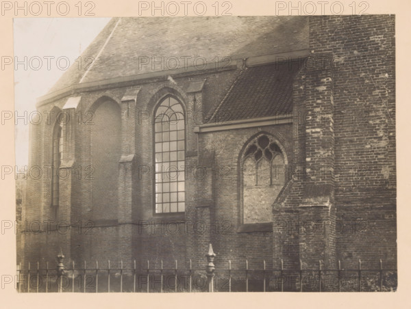 North side of the choir of the Old Church in Naaldwijk, photograph, annotation: 'North side of the choir of the Dutch Reformed Church. May 1910, Naaldwijk', annotation: 'Jz. Sweerts, 1620-1673', Naaldwijk, 1910, photographer, Attributed to, paper, pasteboard, gelatin silver print, height 161 mm x width 225 mm, parts of church exterior and annexes: choir, A sepia-toned photograph of a brick building façade showing steep roof tiles and textured brickwork, with pointed arched window openings and one tall multi-paned window with an arched top, a smaller arched window area with tracery and a boarded or filled interior, buttresses and recessed arches in the brick wall, a sloped section of tiled roof meeting the wall, weathering and patches on the bricks, and an iron fence with vertical posts and decorative finials visible along the bottom edge.