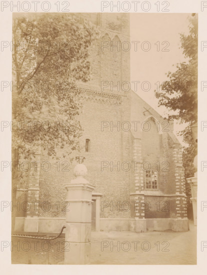 View of the Reformed Church of Noordwijk-Binnen, photograph, annotation: 'Noordwijk-binnen (ZH), NH church, July 1901', annotation: 'NH Church, July 1901, Noordwijk-Binnen', Noordwijk-Binnen, 1901, photographer, Noordwijk, Attributed to, paper, pasteboard, albumen print, height 231 mm x width 170 mm, church (exterior), A sepia-toned photograph showing a brick building with decorative brick patterns near the top and a large arched opening partially filled in, flanked by buttresses with alternating lighter and darker stone blocks, a tall narrow window with multiple panes set into the wall, and a pedestal topped by a rounded stone ornament in the foreground; leafy tree branches frame the left and right sides of the image, and a section of a low wall and paved ground are visible at the bottom.