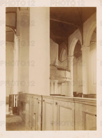 Interior of the Reformed Church of Noordwijk-Binnen, photograph, annotation: 'NH Church, interior, July 1901, Noordwijk-Binnen', annotation: 'Noordwijk-binnen (ZH), NH church, July 1901', Noordwijk-Binnen, 1901, photographer, Noordwijk, Attributed to, paper, pasteboard, albumen print, height 230 mm x width 169 mm, interior of church, A sepia-toned interior view showing tall rounded columns and arches, a high ceiling with visible beams, a wooden balcony with ornate railing partly draped in fabric, a paneled wooden partition or pews running along the foreground with small doors and metal fixtures, a section of a stair or railing at the left, stone or tiled flooring, and soft diffused light coming through tall arched windows.