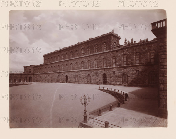 Palazzo Pitti, eretto sui disegni di Brunellesco nel 1440, Palazzo Pitti in Florence, Italy, Firenze, photograph, inscription: 'Firenze, Pitti', number: '36', number: '3037.', photographer: Giacomo Brogi (possibly), Florence, publisher: attributed to Edizioni Brogi, 1864 - 1881, Tuscany, Attributed to, pasteboard, paper, albumen print, height 317 mm x width 445 mm, square, circus, palace, A wide courtyard bordered by a large stone building with a rusticated façade and repeated arched windows on multiple levels, a curved raised walkway with short posts and railings running along the right side, an ornate lamppost with multiple lanterns near the foreground, a paved area leading toward an arched entrance in the building, a section of colonnaded arches visible on the left, decorative balustrades along the roofline, a cloudy sky overhead, and stone chimneys and roof structures visible above the building.