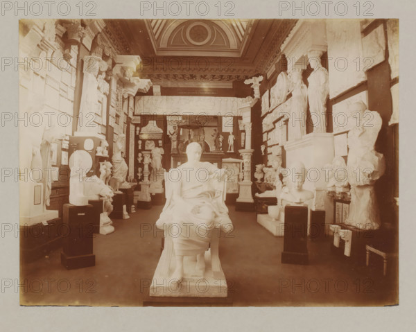 View of a hall with plaster sculptures, album page, photograph, This album page is part of a cover with 6 pages., collector's mark: 'Museum van Kunst-Nijverheid Haarlem', workshop stamp: 'J. Pörtzgen, Photograaf, Koningstraat 38, Haarlem', photographer: Johann Pörtzgen, c. 1890 - c. 1900, Johann Pörtzgen, photographer, paper, pasteboard, daylight collodion silver print, height 166 mm x width 221 mm, sculpture, A large, sepia-toned gallery room filled with many classical white sculptures and reliefs arranged on pedestals and along the walls, with a central seated draped figure in the foreground facing away, flanked by standing statues, busts, and sculpted heads; decorative architectural elements such as columns, capitals, and carved friezes line the upper walls and a coffered ceiling with a circular skylight is visible above, while various plaster casts and sculptural fragments occupy the floor and shelving throughout the space.