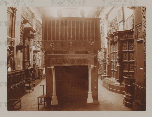 View of a hall with a fireplace and reliefs, album page, photograph, This album page is part of a cover with 6 pages., collector's mark: 'Museum van Kunst-Nijverheid Haarlem', workshop stamp: 'J. Pörtzgen, Photograaf, Koningstraat 38, Haarlem', photographer: Johann Pörtzgen, c. 1890 - c. 1900, Johann Pörtzgen, photographer, paper, pasteboard, daylight collodion silver print, height 166 mm x width 221 mm, open hearth, fire-place, ornamenting, decorating, applied arts, A sepia-toned interior scene showing a room filled with carved wooden furniture and decorative panels, featuring a tall ornate wooden structure with turned balusters and a carved frieze, three small statues positioned on a raised ledge, an elaborately carved mantel with floral and circular motifs supported by slender columns, rows of intricately detailed panels and pilasters lining the walls, several small chairs and stools on a wooden floor, sculpted reliefs and framed carved plaques mounted on the walls, and various orna...