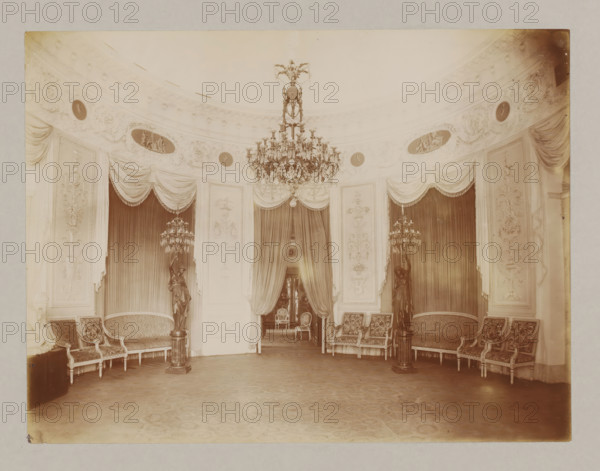 Interior with chandelier and candlesticks, album page, photograph, This album page is part of a cover with 6 pages., workshop stamp: 'J. Pörtzgen, Photographer, Koningstraat 38, Haarlem', collector's mark: 'Museum van Kunst-Nijverheid Haarlem', photographer: Johann Pörtzgen, c. 1890 - c. 1900, Johann Pörtzgen, photographer, paper, pasteboard, daylight collodion silver print, height 166 mm x width 220 mm, chandelier, candelabrum, interior of the house, chair, A large ornate room with a curved decorated cornice and draped valances above tall curtains, a central crystal chandelier hanging from the ceiling, pairs of upholstered settees and chairs arranged along the walls, two dark statues on pedestals holding multi-arm candle fixtures flanking a doorway, decorative wall panels with sculpted motifs, an open doorway revealing another room with chairs, and a patterned floor with visible wear.
