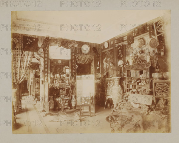 Interior, album page, photograph, This album page is part of a cover with 6 pages., collector's mark: 'Museum van Kunst-Nijverheid Haarlem', workshop stamp: 'J. Pörtzgen, Photograaf, Koningstraat 38, Haarlem', photographer: Johann Pörtzgen, c. 1890 - c. 1900, Johann Pörtzgen, photographer, paper, pasteboard, daylight collodion silver print, height 166 mm x width 220 mm, interior of the house, mirror, decorations (interior of the house), A sepia-toned interior filled with ornate decorative objects and furniture, including large vases, carved wooden chairs, a small round table, and shelves holding figurines and bowls; patterned curtains hang at openings and tapestries or framed panels cover walls, with circular plates and woven items displayed; a carved ornamental screen and a draped fabric panel appear near the center, while smaller stools and cabinets with intricate detailing occupy the floor space; various hanging ornaments, a cluster of sticks or poles, and a textured rug or animal h...