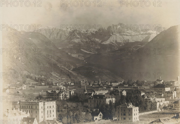 Landscape with mountains and a city, album page, photograph, This photo is part of an album., annotation: 'From Breitner's studio', 1880 - 1920, photographer, paper, pasteboard, gelatin silver print, developed gelatin silver print, height 129 mm x width 180 mm, Photo is loose in the front of the album., city-view, and landscape with man-made constructions, mountains, A panoramic view shows a town with clustered buildings and multi-story houses in the foreground, some with pitched roofs and decorative facades, interspersed with trees and fenced yards; further back a denser area of rooftops and church spires rises along a gentle slope leading into terraced fields and vineyards on the lower mountain slopes; beyond those the terrain steepens into dark, forested hills that give way to a dramatic range of jagged, snow-capped mountains under a hazy sky, with soft gradients of shadow and light across the valleys.