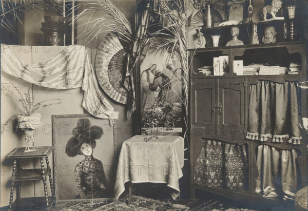 Interior with a cupboard, sculptures and a painting, album page, photograph, This photo is part of an album., annotation: 'From Breitner's studio', 1880 - 1920, photographer, paper, pasteboard, gelatin silver print, developed gelatin silver print, height 129 mm x width 180 mm, Photo is loose at the front of the album., sculpture, picture, painting, interior of the house, furniture to put things on, furniture for storage, A room corner filled with decorative objects and furnishings, including a wooden cabinet with shelves displaying vases, busts, a small figurine, folded textiles, and stacked papers, curtains with fringe hanging from some openings, and patterned fabric visible behind lower openings; a table covered with a patterned cloth supporting a small plant and a tray with glassware; a large framed portrait of a woman wearing a hat leaning against the wall near a small turned-leg side table that holds a pedestal planter with a fern and a ruffled fabric cover; a draped silk or satin...