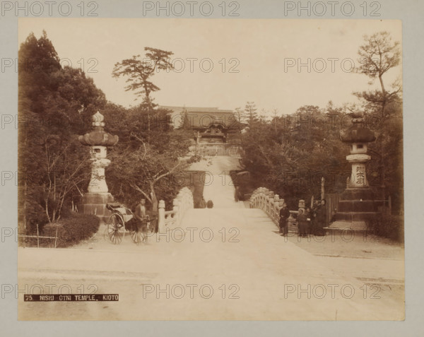75. Nishi-Otni Temple.-Kioto, Nishi-Otani Temple, Kyoto, photograph, number: '7.', c. 1895 - c. 1915, photographer, Kinki, paper, pasteboard, photo paper on cardboard, height 210 mm x width 270 mm, height 243 mm x width 329 mm, A wide view of a stone bridge leading toward a distant temple gate framed by tall trees on both sides, with ornate stone lanterns atop pedestals flanking the bridge entrance, a rickshaw parked near the left lantern with a man standing beside it, a group of people standing near the right lantern, stone railings lining the bridge, trees and foliage surrounding the pathway, and a roofed temple structure visible beyond the top of the steps.