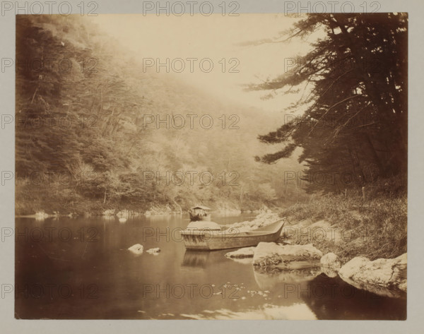 River of Hozu, Kyoto, photograph, A boat is moored on the right, a boat with a roof is sailing in the background., annotation: '10. Eind der Hodzu-rapids', c. 1895 - c. 1915, photographer, Kinki, paper, pasteboard, albumen print, albumendruk, height 212 mm x width 271 mm, height 243 mm x width 329 mm, A sepia-toned riverside scene showing a calm stretch of water bordered by rocky banks and dense trees, with a small wooden rowboat pulled up on the near shore and another covered boat floating further out, a larger tree with overhanging branches occupying the right side while the left side opens to a hillside of foliage fading into mist, reflected shapes visible on the water surface and scattered rocks breaking the waterline.