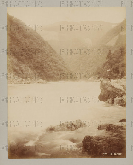Rapids in Uji River, Uji, 705 Uji Rapids, photograph, number: '11.', c. 1895 - c. 1915, photographer, Kinki, Kyoto, paper, pasteboard, albumen print, height 271 mm x width 212 mm, height 329 mm x width 243 mm, A sepia-toned scene showing a river flowing through a narrow valley flanked by steep, tree-covered hills, with misty distant ridges; rocks and turbulent water occupy the foreground and midriver area, and a person wearing a conical hat stands on a rocky outcrop at the river edge holding a long pole with a small container beside them.