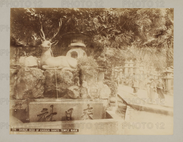 Bronze deer of the Kasuga Shinto Temple, Nara, 776 Bronze deer of Kasuga Shinto Temple Nara, photograph, number: '14.', c. 1895 - c. 1915, photographer, Kinki, paper, pasteboard, albumen print, albumendruk, height 209 mm x width 270 mm, height 243 mm x width 329 mm, A sepia-toned photograph showing a large reclining deer statue with prominent antlers positioned on a rocky ledge, water appearing to flow from its mouth into a stone basin below; behind the statue is a rounded lantern structure partially obscured by foliage and trees with dense leafy branches; to the right a walkway runs alongside stone lanterns and stone posts, and several people wearing traditional clothing and hats are walking or standing along the path, with some closer to the foreground and others further back among the stone structures.