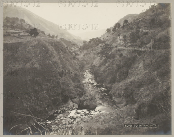 River between mountains, Kanagawa, Road to Miyanoshita, photograph, number: '16.', c. 1895 - c. 1915, photographer, Honshu, paper, pasteboard, gelatin silver print, developing gelatin silver print, height 229 mm x width 292 mm, height 243 mm x width 329 mm, A grayscale photograph of a narrow river flowing between steep, tree-covered hills with rocky banks, showing water cascading over boulders and winding toward a distant, misty valley; vegetation and scattered trees cover the slopes on both sides, with some bare branches and exposed rock faces visible, and utility poles with crossarms and wires stand on a hillside at one side of the scene.