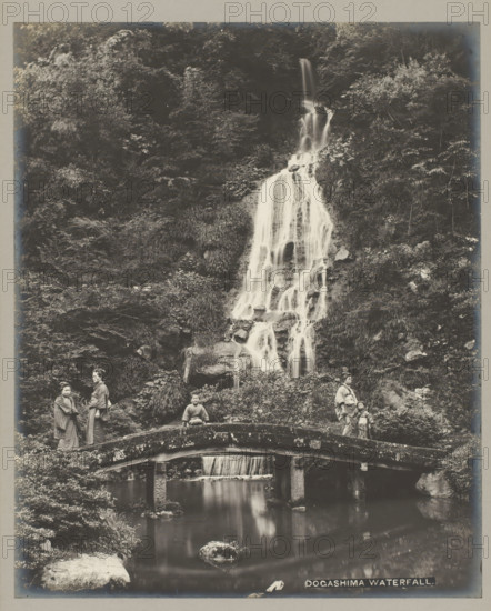 Dogashima waterfall., Dogashima waterfall, Kanagawa-Ken, photograph, number: '19.', c. 1895 - c. 1915, photographer, Kanto, paper, pasteboard, gelatin silver print, developing gelatin silver print, height 283 mm x width 229 mm, height 329 mm x width 243 mm, A tall, multi-tiered waterfall cascades down a rocky, vegetated hillside into a pool below, with water flowing over rocks and through grassy vegetation; a stone arched bridge spans the pool in the foreground, its surface mottled and textured, and beneath the bridge a smaller fall feeds the reflective water; on the bridge and near it stand several people wearing traditional garments, including adults and children, positioned at different points along the bridge and banks, with one person seated on the bridge; dense foliage and trees cover the surrounding slopes, and rocks and plants frame the scene on both sides.