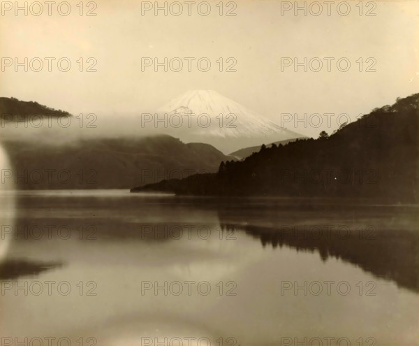 Fuji seen from a distance, Japan, photograph, annotation: '23. Fuji', c. 1895 - c. 1915, photographer, paper, pasteboard, gelatin silver print, developing gelatin silver print, height 229 mm x width 281 mm, height 243 mm x width 329 mm, A wide, misty landscape with a snow-capped conical mountain in the distance partially veiled by low clouds, darker forested hills on either side framing the mountain, a calm reflective body of water in the foreground mirroring the hills and sky, and a soft, pale sky above.