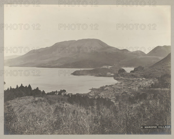 8 Hakone village., View of village and Hakone Lake, Hakone, photograph, number: '24.', c. 1895 - c. 1915, photographer, paper, pasteboard, gelatin silver print, developing gelatin silver print, height 222 mm x width 287 mm, height 243 mm x width 329 mm, A wide grayscale landscape showing a large calm body of water bordered by gently sloping hills and a prominent rounded mountain in the background, with clusters of trees on the nearer slopes and a spread of small buildings with pitched roofs along the shoreline, a narrow peninsula jutting into the water with a larger light-colored building near its tip, and foreground grasses and shrubs covering the hillside from which the scene is viewed.