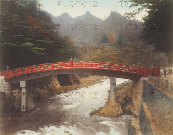 Sacred bridge, Japan, photograph, annotation: '27. Sacred bridge.', c. 1895 - c. 1915, photographer, paper, pasteboard, salted paper print, Salt print, colored., height 208 mm x width 267 mm, height 243 mm x width 329 mm, A red arched wooden bridge spans a flowing river, supported by stone and concrete piers, with carved railings and decorative finials along its length; rocky banks and a stone retaining wall border the river, and a pathway runs beside the wall. Dense trees and foliage frame both sides of the scene, with a mix of dark green and autumnal leaves visible, and distant misty mountains rise in the background under a pale sky. Two small human figures stand near the far end of the bridge, partially obscured, and scattered rocks and shallow rapids appear in the river beneath the bridge.