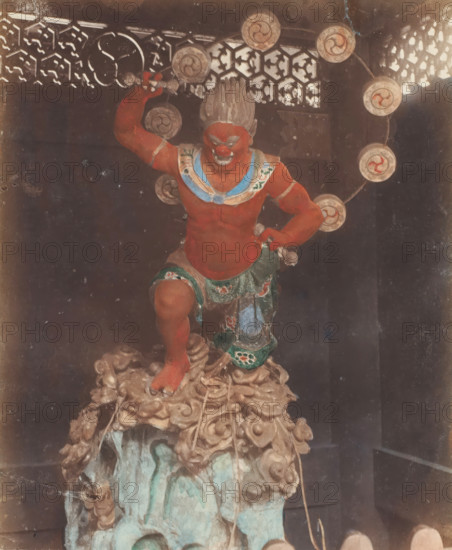 B 85 The Thunder God of Buddest Temple Nikko, God of Thunder at Buddhist temple, Nikko, photograph, number: '31.', c. 1895 - c. 1915, photographer, Kanto, paper, pasteboard, salted paper print, Salt print, colored., height 253 mm x width 201 mm, height 329 mm x width 243 mm, A painted statue of a muscular red-skinned figure stands on a carved base that resembles swirling clouds and rock, the figure posed with one leg raised and one arm bent upward holding a small object, wearing a decorative skirt with green and patterned details and a blue and white collar, the face showing a fierce expression with prominent brows and a wide mouth and hair styled upward, a circular halo of disks with spiral motifs arches behind the figure, an ornate lattice screen with geometric cutouts is visible in the upper background, and the image shows signs of age with scratches and discoloration.