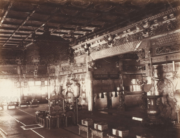 B 3 A front hall of Buddest Temple Nikko., Hall in the front of Buddhist temple, Nikko, photograph, number: '3.3.', c. 1895 - c. 1915, photographer, Kanto, paper, pasteboard, salted paper print, Zoutdruk, height 210 mm x width 265 mm, height 243 mm x width 329 mm, An ornate interior space with a decorated coffered ceiling and carved beams, large hanging lanterns and decorative cords suspended from above, intricately carved wooden pillars and beams with repeating geometric and floral motifs along the upper walls, rows of low wooden stools and small ornate boxed platforms arranged on the floor, tall ornamental vases and stands with sculpted details positioned near the front, carved guardian figures flanking the sides with curled manes and stylized faces, hanging decorative garlands and plaques along the walls, patterned floor coverings and runners defining walkways, and light coming through windows that casts shadows across the furnishings.