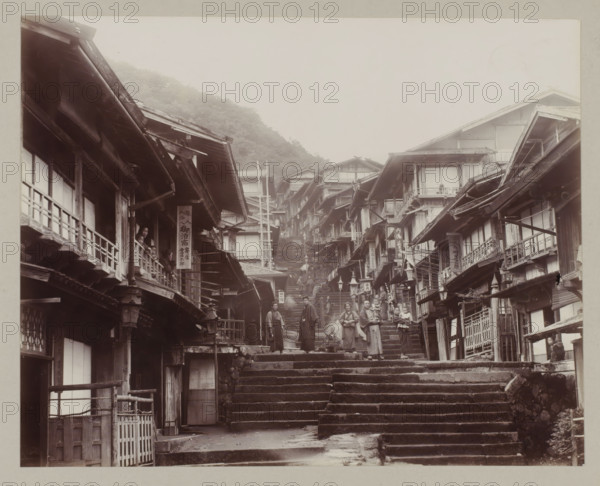 Street in Ikaho with steps, photograph, annotation: '42. Ikao.', Ikaho, c. 1895 - c. 1915, photographer, Kanto, paper, pasteboard, daylight collodion silver print, daylight collodion silver print on photo paper on cardboard, height 201 mm x width 260 mm, height 243 mm x width 329 mm, A narrow stone stairway ascends between closely packed wooden buildings with overhanging eaves and balconies, the façades showing exposed beams and lattice windows; people in traditional clothing stand and walk on the steps and on the balconies, some holding children, others gathered in small groups; wooden signboards hang from several buildings but their content is not described; the street slopes upward toward a misty hillside with trees visible in the background, and lanterns and railings line portions of the stairs and building fronts.