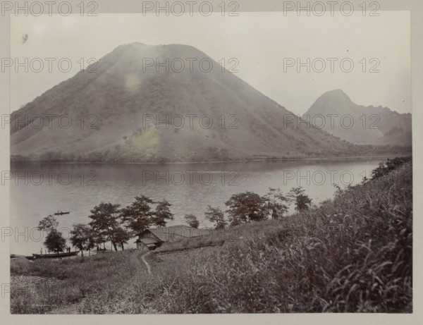 More from Haruna, photograph, annotation: '44. Haruna lake.', Haruna, c. 1895 - c. 1915, photographer, Kanto, paper, pasteboard, daylight collodion silver print, daylight collodion silver print on photo paper on cardboard, height 210 mm x width 280 mm, height 243 mm x width 329 mm, A wide lake fills the middle ground, its surface smooth and reflective, bordered by a shoreline with scattered trees and grassy slopes; a large rounded mountain rises steeply from the far shore, covered in textured vegetation, and a smaller jagged peak appears further back to the right; in the foreground a hillside with tall grasses slopes down toward a rustic wooden building with a pitched roof nestled among trees near the water, and a narrow footpath leads down the slope; small boats float on the lake, one with a person standing, and leafy trees frame the lower edge of the scene.