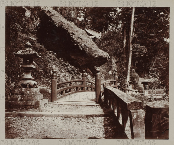 Entrance of the Haruna temple, photograph, annotation: '46. Entrance to Haruna temple.', Haruna, c. 1895 - c. 1915, photographer, Kanto, paper, pasteboard, daylight collodion silver print, daylight collodion silver print on photo paper on cardboard, height 217 mm x width 268 mm, height 243 mm x width 329 mm, A stone lantern stands on a stepped base to the left of a gently arched wooden bridge with rounded posts topped by bulbous finials, the bridge leading toward a rocky hillside and dense trees; a large overhanging rock looms above the scene, and a worn stone railing runs along the right side of the path, with scattered pebbles and leaves on the ground and a set of stone steps and small structures partially visible among the trees in the background.