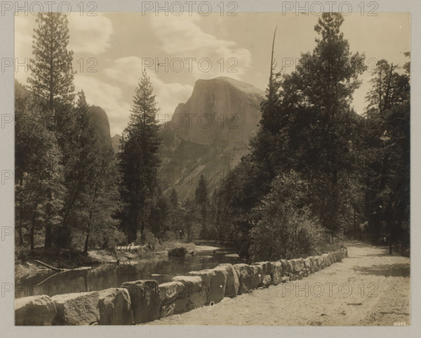 Yosemite Valley, photograph, Bottom right reads: 51083., annotation: '3. Yosemete Valley', California, c. 1895 - c. 1915, photographer, North America, paper, pasteboard, gelatin silver print, developing gelatin silver print, height 189 mm x width 239 mm, height 243 mm x width 329 mm, A sepia-toned landscape photograph showing a rocky mountain with a rounded top framed by tall conifer trees and dense foliage, a calm river running through the foreground with reflections of nearby trees, a low stone wall lining a dirt path beside the river, scattered large boulders and logs near the water, and a lamp post partially visible among the trees along the path.