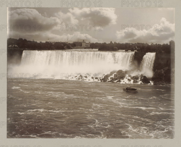 Niagara waterfall, United States, Canada, photograph, Niagara waterfall seen from the front, on the right a boat sails., number: '8.', Niagara Falls (North America), c. 1895 - c. 1915, photographer, North America, paper, pasteboard, gelatin silver print, developing gelatin silver print, height 182 mm x width 239 mm, height 243 mm x width 329 mm, A wide view of a large waterfall with water cascading over a cliff into a misty, rocky plunge below, a river with rippling, foamy water in the foreground, a small boat on the river near the waterfall, rocky outcrops and spray at the waterfall base, a tree-lined shoreline and buildings visible on the far bank above the falls, and a sky filled with large, billowing clouds.