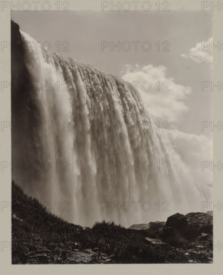 Niagara waterfall seen from below, United States, Canada, photograph, number: '9.', Niagara Falls (North America), c. 1895 - c. 1915, photographer, North America, paper, pasteboard, gelatin silver print, developing gelatin silver print, height 239 mm x width 189 mm, height 329 mm x width 243 mm, A large curtain of water cascades over a cliff, filling the left and central parts of the image with rushing, misty flows; the water appears textured with streams and softer, blurred areas where it falls. A vertical dark rock face is visible at the top left where the water begins to spill over. Clouds occupy the upper right portion of the scene, appearing soft and diffuse against a pale sky. In the lower foreground there is rocky ground with patches of grass and low vegetation, and a few larger rocks along the bottom edge. The overall image is monochromatic with high contrast between the bright falling water and the darker rocks and vegetation.